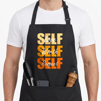 Self x apron - Stacked orange SELF text with white Love Worth Respect script overlay — shown on a black cotton canvas apron against a white background