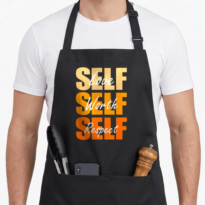 Self x apron - Stacked orange SELF text with white Love Worth Respect script overlay — shown on a black cotton canvas apron against a white background