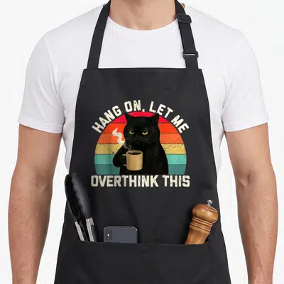 Hang coffee aprons - Black cat holding mug under arched text over striped semi-circle sunset — shown on a black cotton canvas apron against a white background