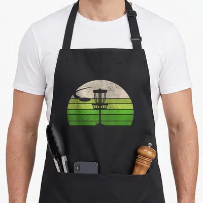 Frisbee ultimate apron - Retro disc golf basket and flying disc against striped green and beige sun background — shown on a black cotton canvas apron against a white background