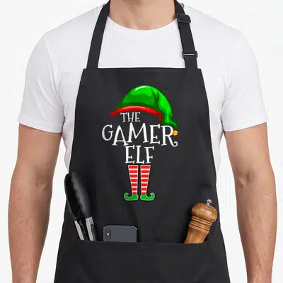Gamer christmas elf apron - THE GAMER ELF text between green hat and striped legs — shown on a black cotton canvas apron against a white background