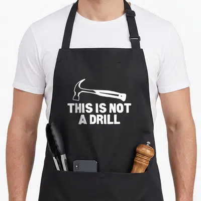 Drill builder apron - White hammer illustration above text THIS IS NOT A DRILL on black fabric — shown on a black cotton canvas apron against a white background