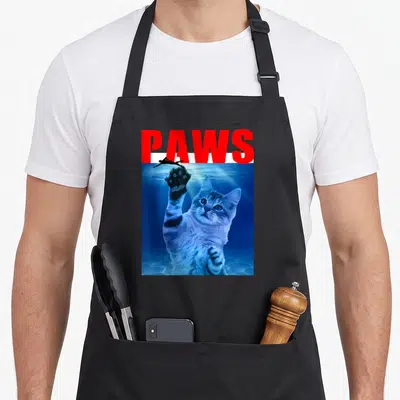 Paw parody cat apron - Red PAWS text above blue underwater cat reaching for black swimmer silhouette — shown on a black cotton canvas apron against a white background