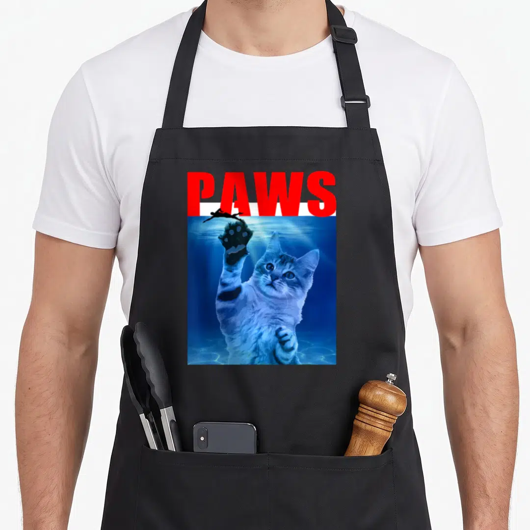 Paw parody cat apron - Red PAWS text above blue underwater cat reaching for black swimmer silhouette — shown on a black cotton canvas apron against a white background