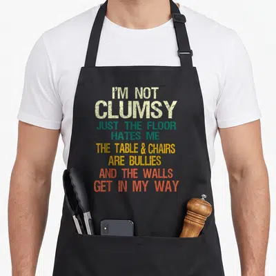 Cute clumsy sarcastic apron - I'M NOT CLUMSY typography in beige, green, yellow, and orange-red stacked text — shown on a black cotton canvas apron against a white background