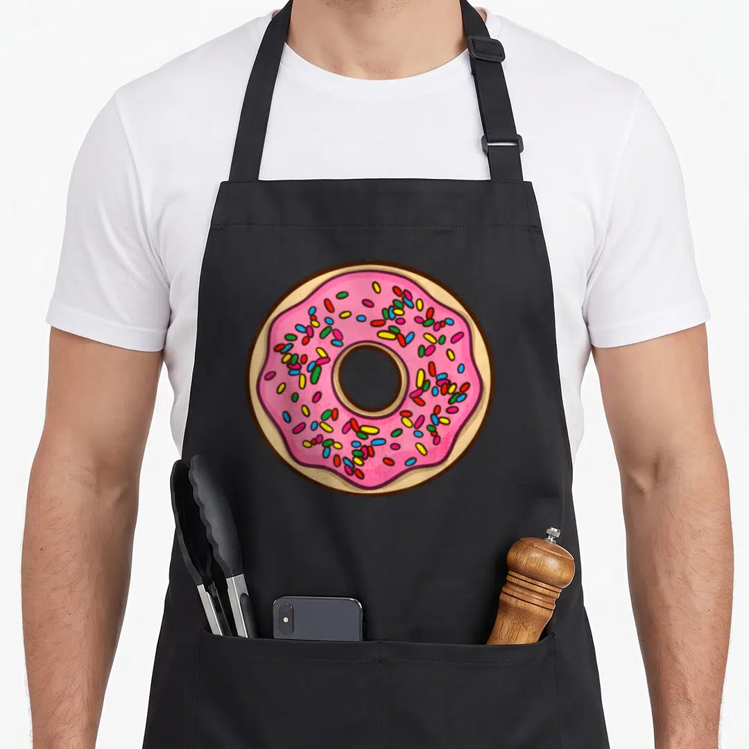 Frying donut apron - Cartoon pink frosted donut with red, blue, yellow, green, and tan sprinkles — shown on a black cotton canvas apron against a white background