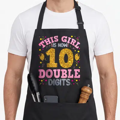 10th birthday apron - Gold number 10 with pink double digits text, balloons, and confetti — shown on a black cotton canvas apron against a white background