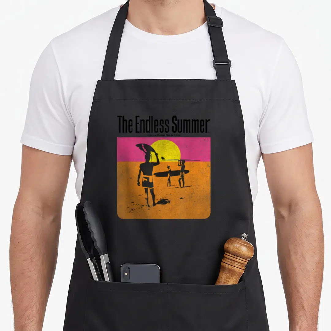 Endless 60s apron - The Endless Summer text above three black surfer silhouettes on orange beach with yellow sun — shown on a black cotton canvas apron against a white background