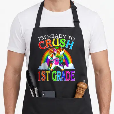 Ready crush unicorn apron - Rainbow cartoon unicorn dabbing with backpack containing apple and pencil under split text — shown on a black cotton canvas apron against a white background
