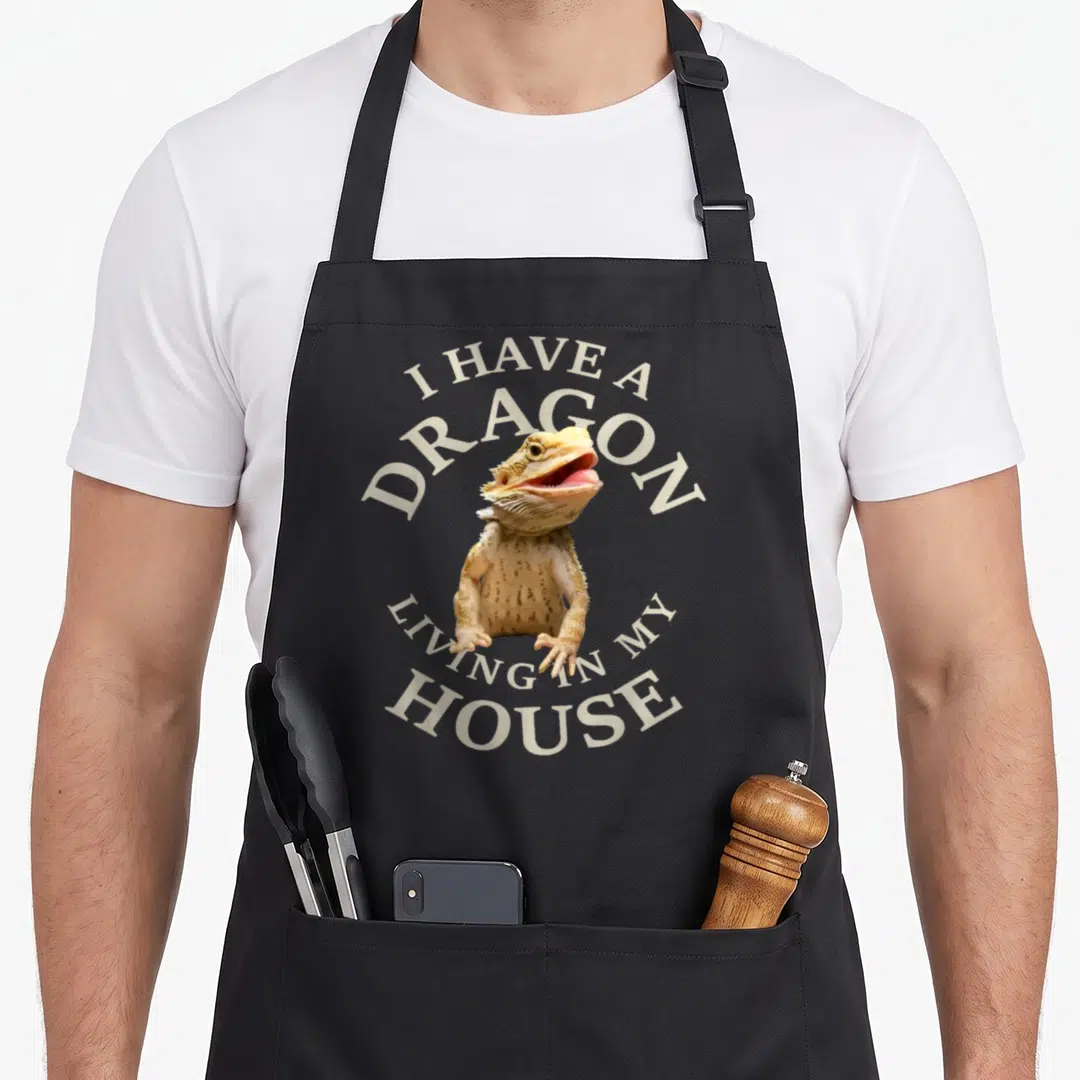 Barbata dragon apron - Bearded dragon photo with circular text I HAVE A DRAGON LIVING IN MY HOUSE in beige and tan — shown on a black cotton canvas apron against a white background
