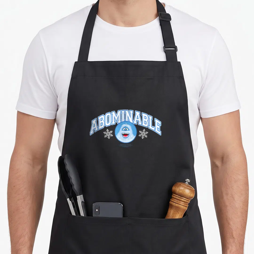 Bumble christmas apron - ABOMINABLE text arched over blue circle with smiling yeti head and two snowflakes — shown on a black cotton canvas apron against a white background