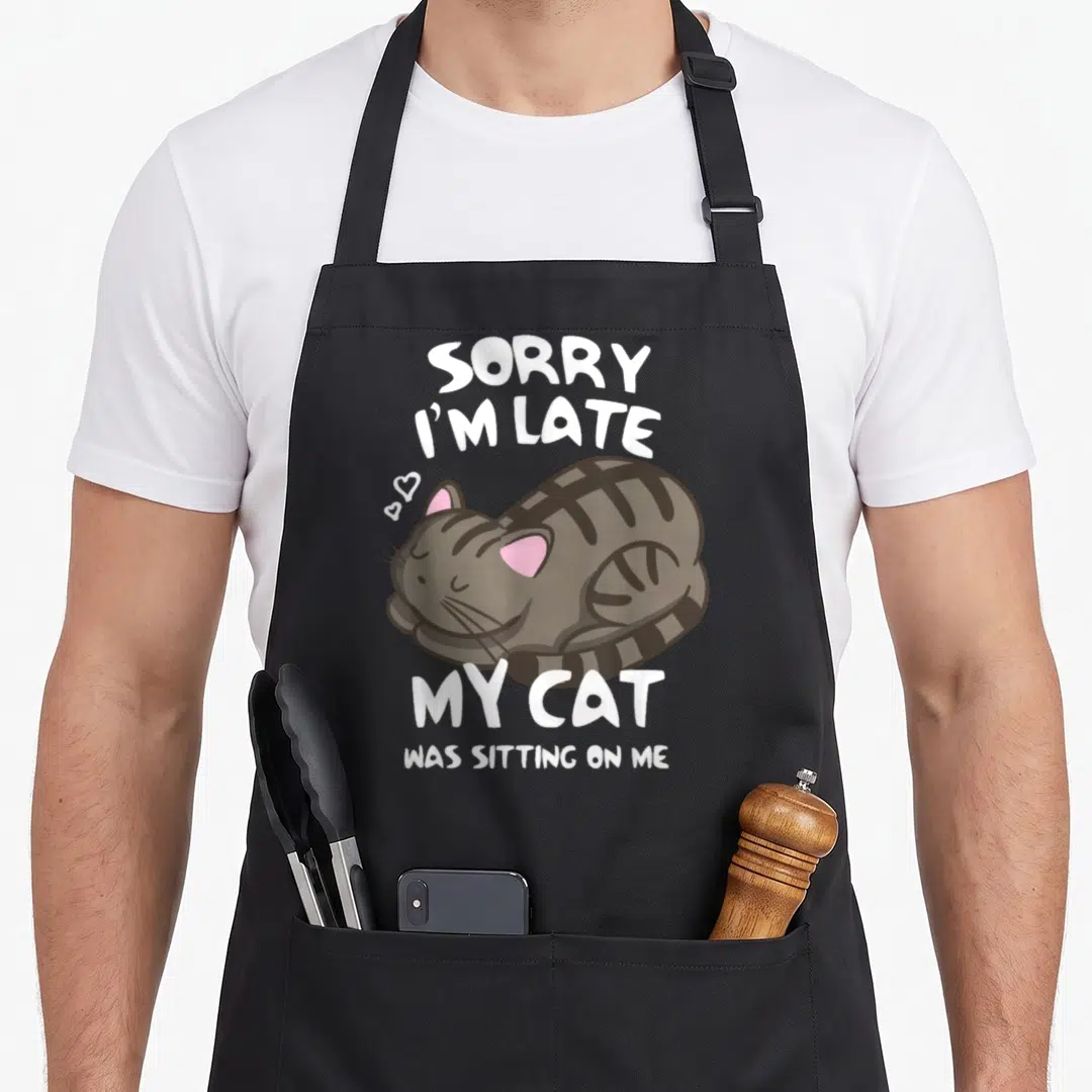 Sitting cat apron - Sleeping grey tabby cat with pink ears and hearts surrounded by sorry I'm late text — shown on a black cotton canvas apron against a white background