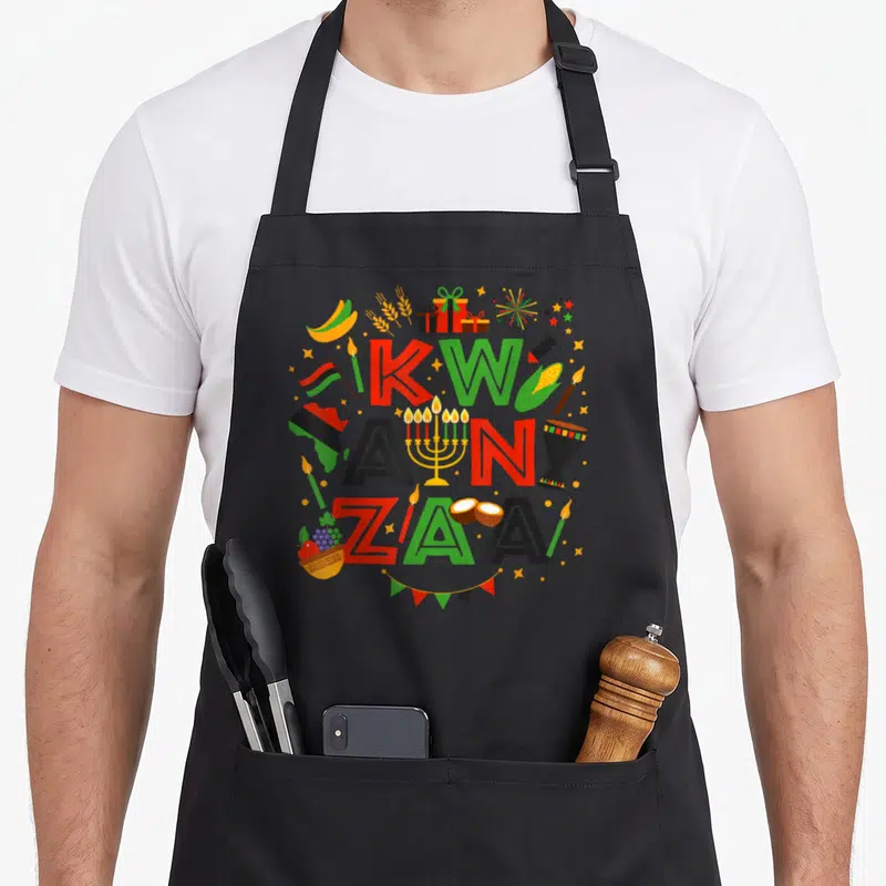 Funny kwanzaa apron - Stacked KWA ANZA ZAA text with corn, drum, flag, and bunting in red green black yellow cartoon — shown on a black cotton canvas apron against a white background