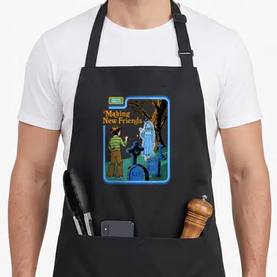 Steven making an apron - Cartoon boy waving at blue ghost in graveyard with SOCIAL SKILLS text — shown on a black cotton canvas apron against a white background