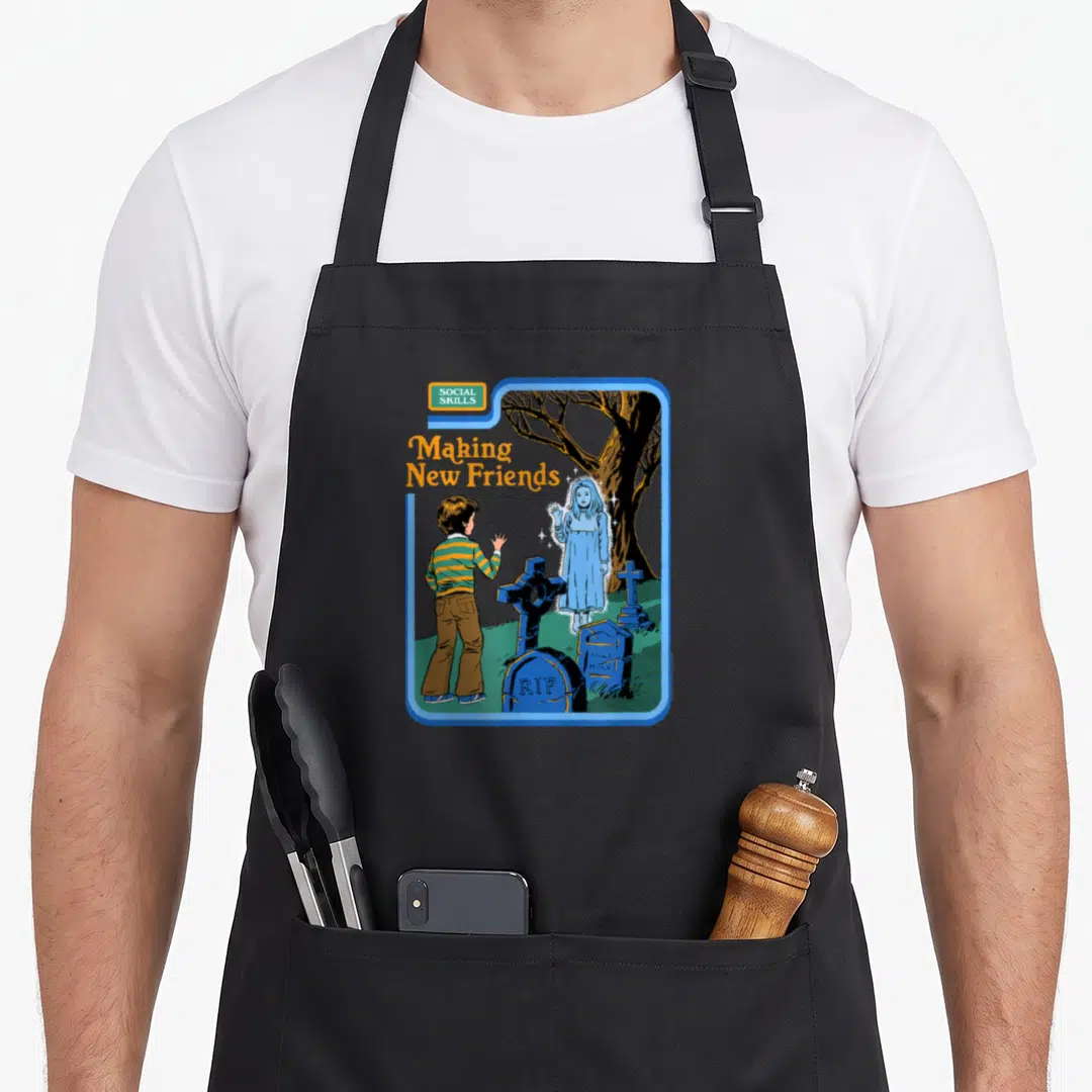 Steven making an apron - Cartoon boy waving at blue ghost in graveyard with SOCIAL SKILLS text — shown on a black cotton canvas apron against a white background