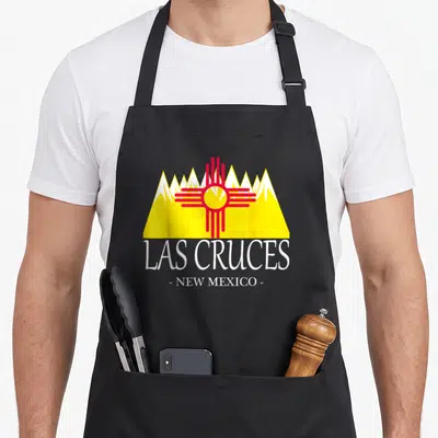 Las cruce mexico apron - Red Zia sun over yellow mountains with white LAS CRUCES text — shown on a black cotton canvas apron against a white background