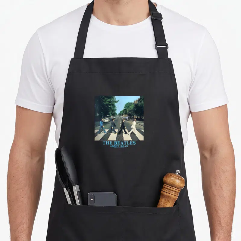 Abbey road apron for women - Square photo of four men walking on crosswalk with blue THE BEATLES ABBEY ROAD text below — shown on a black cotton canvas apron against a white background
