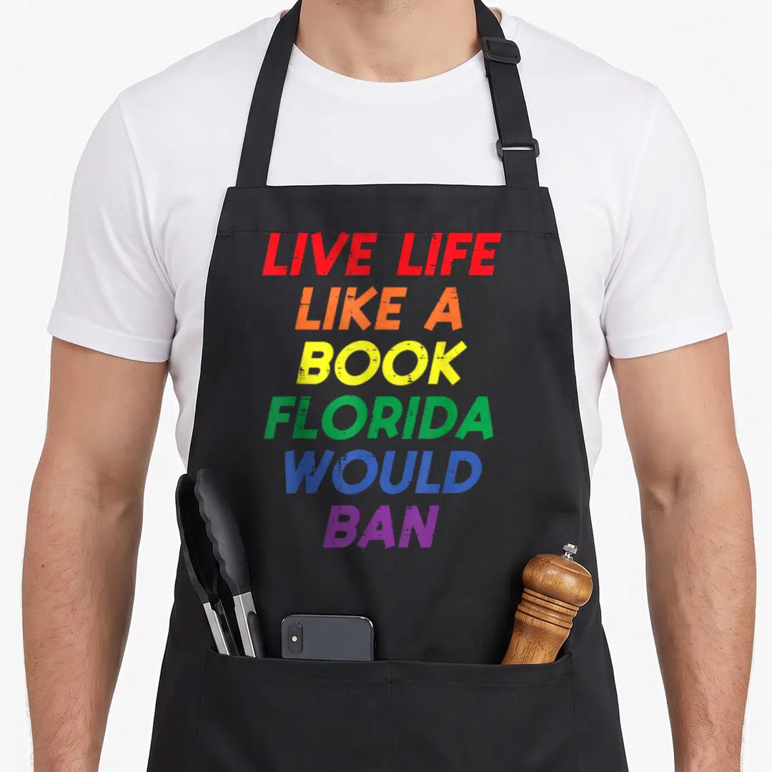 Live gay pride apron - Stacked rainbow text reading LIVE LIFE LIKE A BOOK FLORIDA WOULD BAN — shown on a black cotton canvas apron against a white background