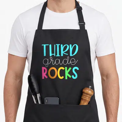 Third art teacher aprons - THIRD grade ROCKS text in blue, white, pink, orange, yellow, and green stacked vertically — shown on a black cotton canvas apron against a white background