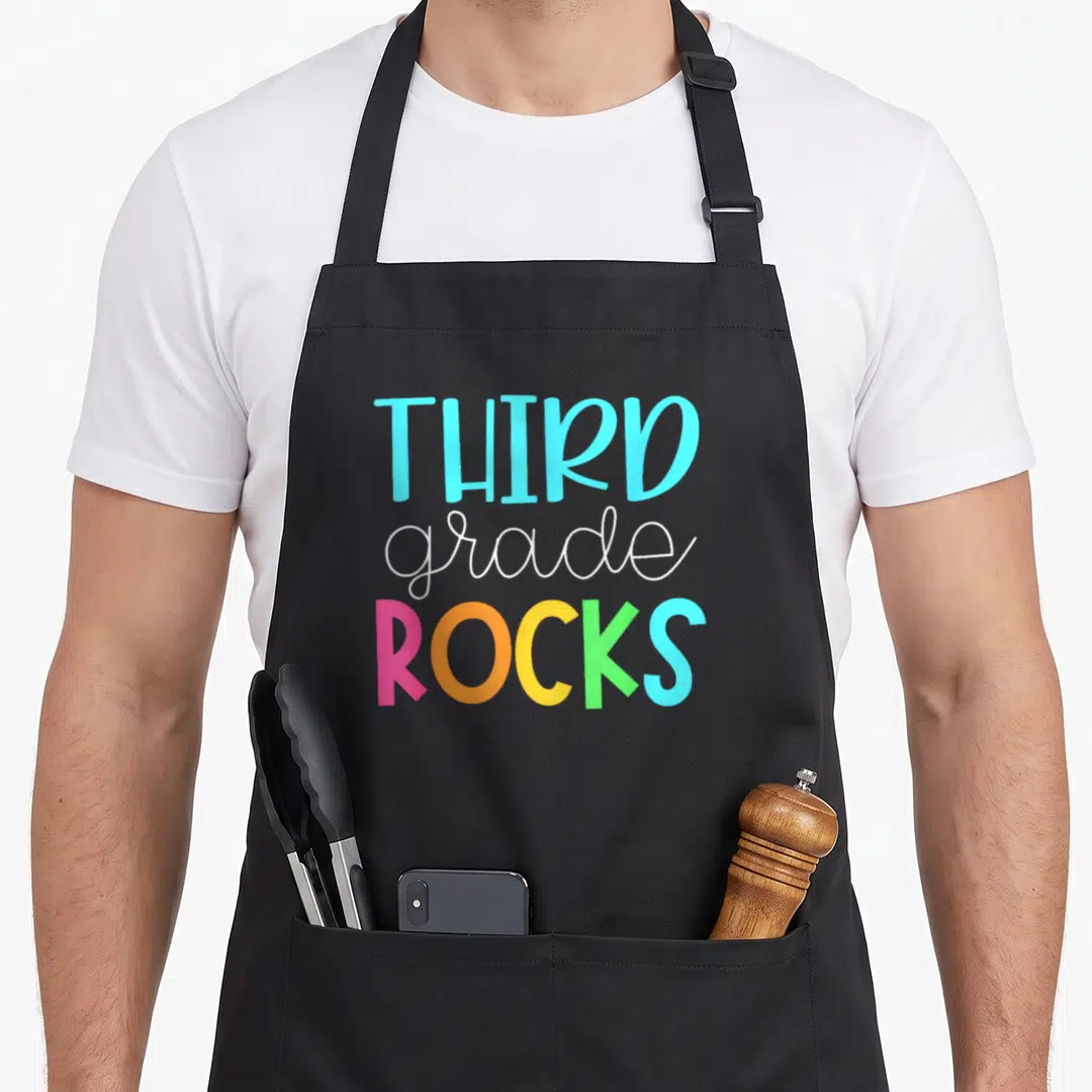 Third art teacher aprons - THIRD grade ROCKS text in blue, white, pink, orange, yellow, and green stacked vertically — shown on a black cotton canvas apron against a white background