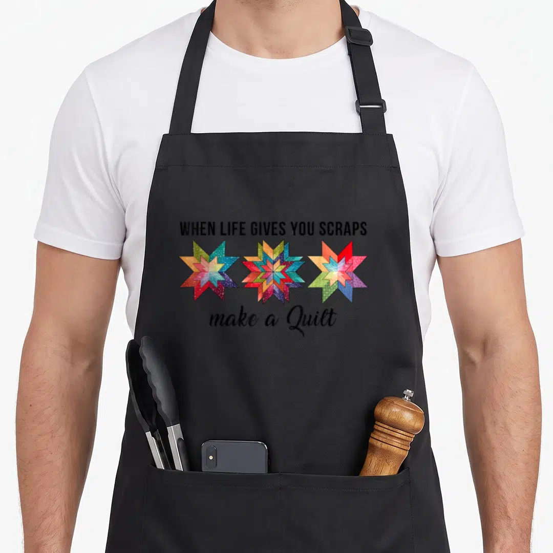Give quilt apron - Three multicolored quilt stars with black text reading WHEN LIFE GIVES YOU SCRAPS make a Quilt — shown on a black cotton canvas apron against a white background