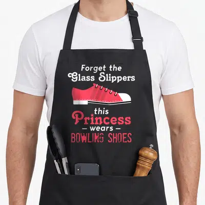 Funny bowling wife apron - Cartoon pink bowling shoe with text forget glass slippers princess wears bowling shoes — shown on a black cotton canvas apron against a white background