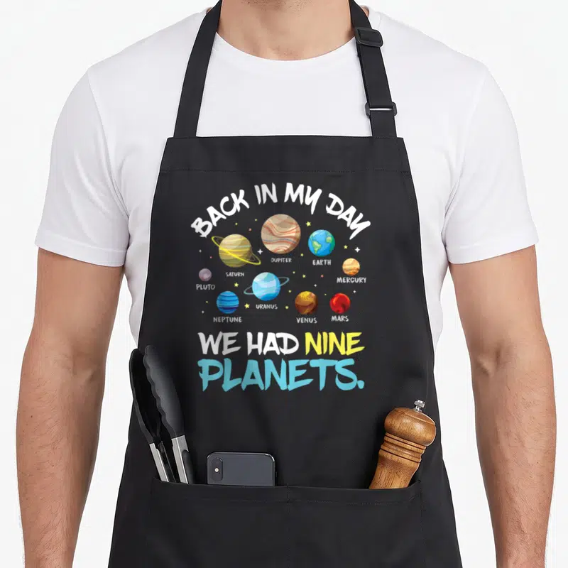 Nine astronaut back apron - Cartoon illustration of nine planets with text Back In My Day We Had Nine Planets — shown on a black cotton canvas apron against a white background