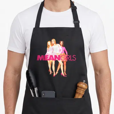 Mean round neck apron - Three women in pink outfits with MEAN GIRLS text across legs — shown on a black cotton canvas apron against a white background
