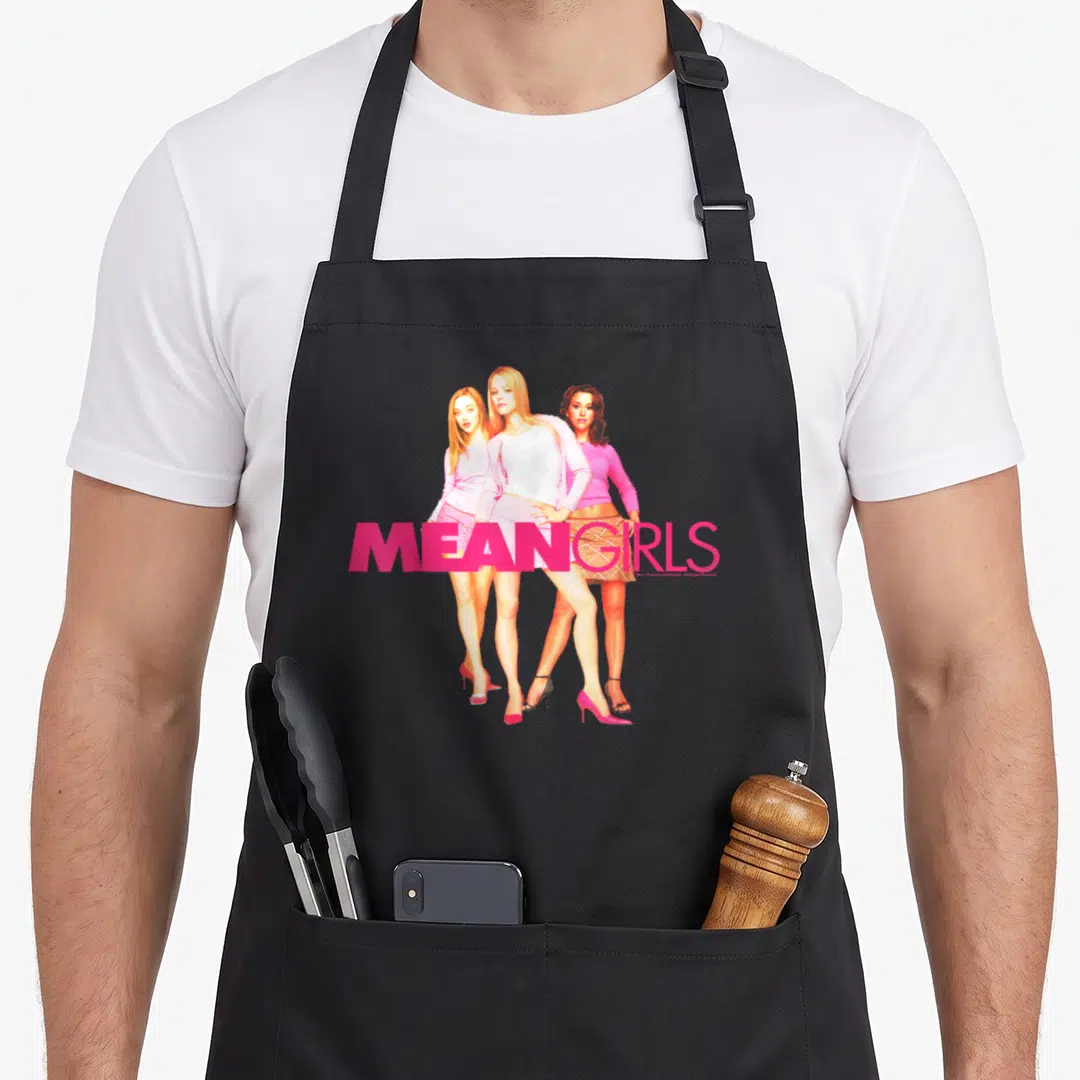 Mean round neck apron - Three women in pink outfits with MEAN GIRLS text across legs — shown on a black cotton canvas apron against a white background