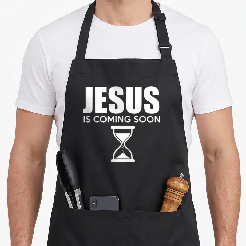 Jesus advent apron - White JESUS IS COMING SOON text above an hourglass icon — shown on a black cotton canvas apron against a white background