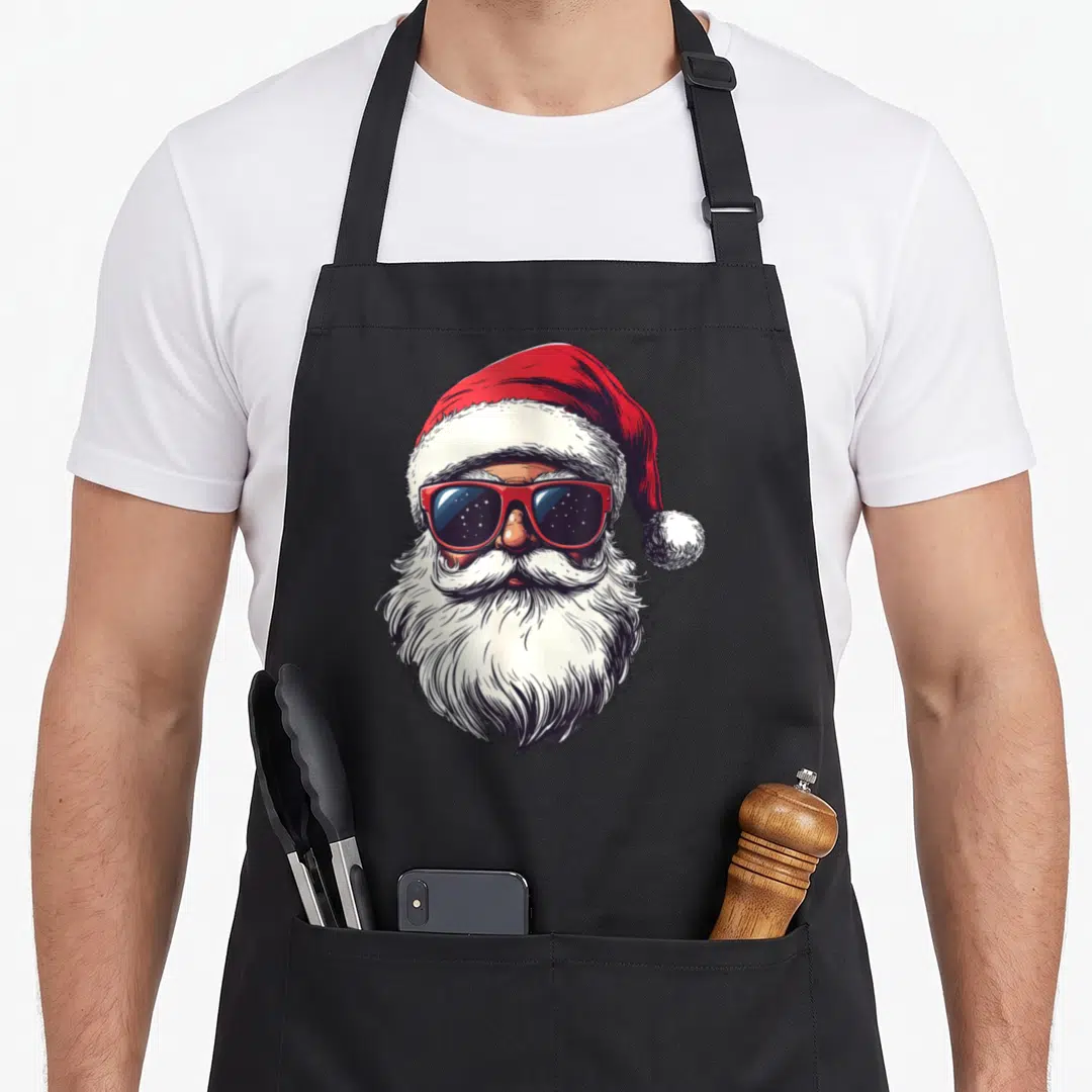 Face santa claus apron - Cartoon Santa head with red sunglasses and white beard on red hat — shown on a black cotton canvas apron against a white background