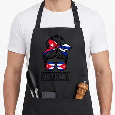 Cubanita messy aprons - Cartoon woman bun with Cuban flag sunglasses and CUBANITA text in black border — shown on a black cotton canvas apron against a white background