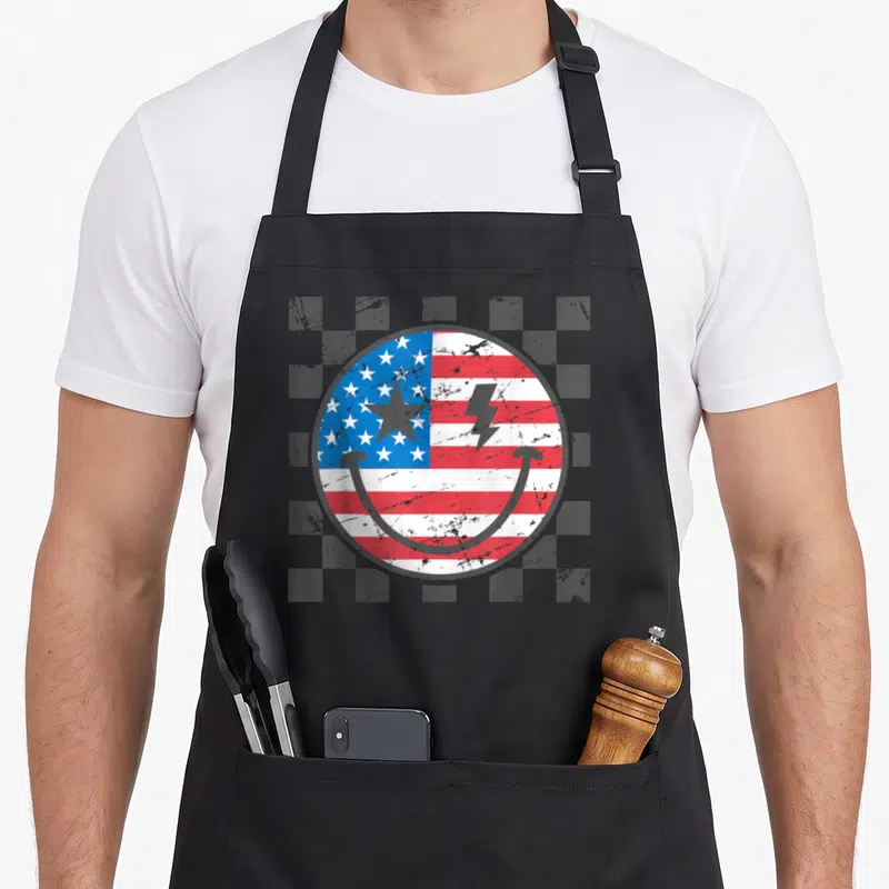 4th face on apron - Distressed smiley face with USA flag pattern and lightning bolt on checkered background — shown on a black cotton canvas apron against a white background