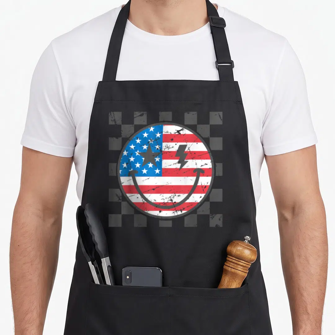 4th face on apron - Distressed smiley face with USA flag pattern and lightning bolt on checkered background — shown on a black cotton canvas apron against a white background