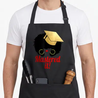 Queen master apron - Minimalist woman silhouette with afro, gold cap, red text saying Mastered it! — shown on a black cotton canvas apron against a white background
