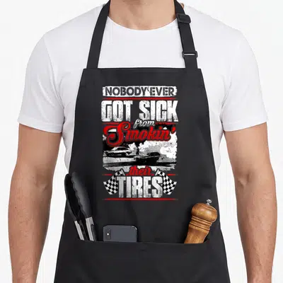 Nobody smoking apron - Stacked text reading nobody ever got sick from smokin their tires above two cars doing a burnout — shown on a black cotton canvas apron against a white background