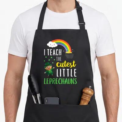 Teach school apron - Rainbow, smiling cloud, pot of gold, leprechaun, and stacked text reading I TEACH THE cutest — shown on a black cotton canvas apron against a white background
