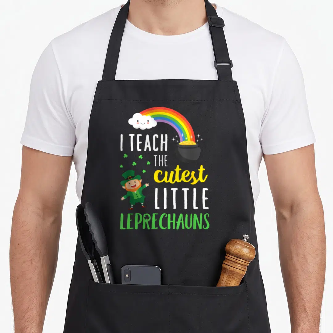 Teach school apron - Rainbow, smiling cloud, pot of gold, leprechaun, and stacked text reading I TEACH THE cutest — shown on a black cotton canvas apron against a white background