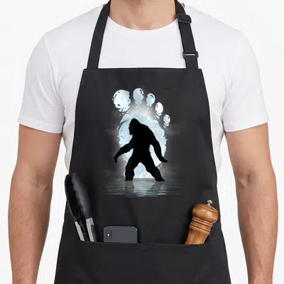 Footprint bigfoot apron - Black Bigfoot silhouette standing before a glowing light blue footprint reflected in water — shown on a black cotton canvas apron against a white background