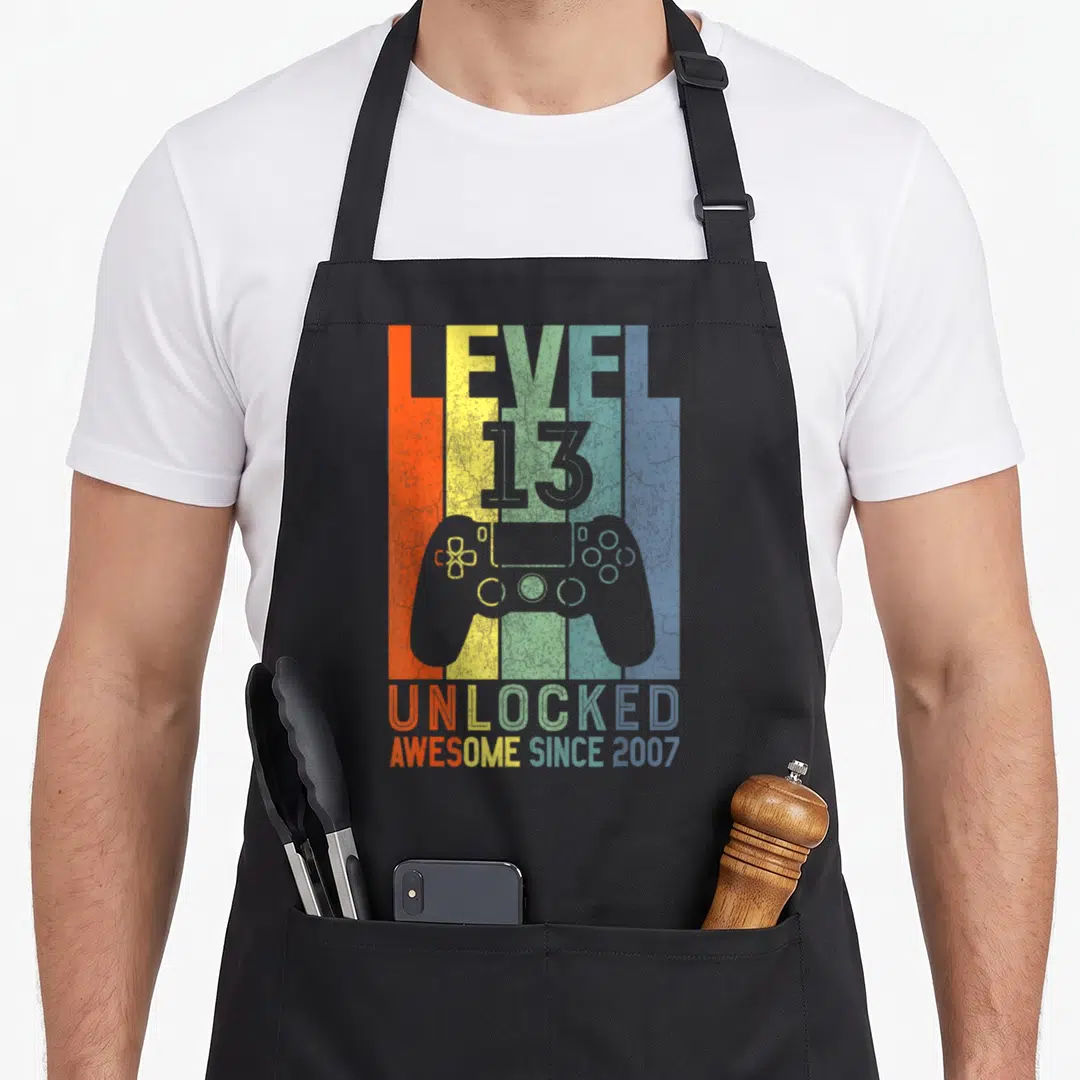 Retro level birthday apron - Game controller over orange yellow green blue stripes with LEVEL 13 UNLOCKED text — shown on a black cotton canvas apron against a white background