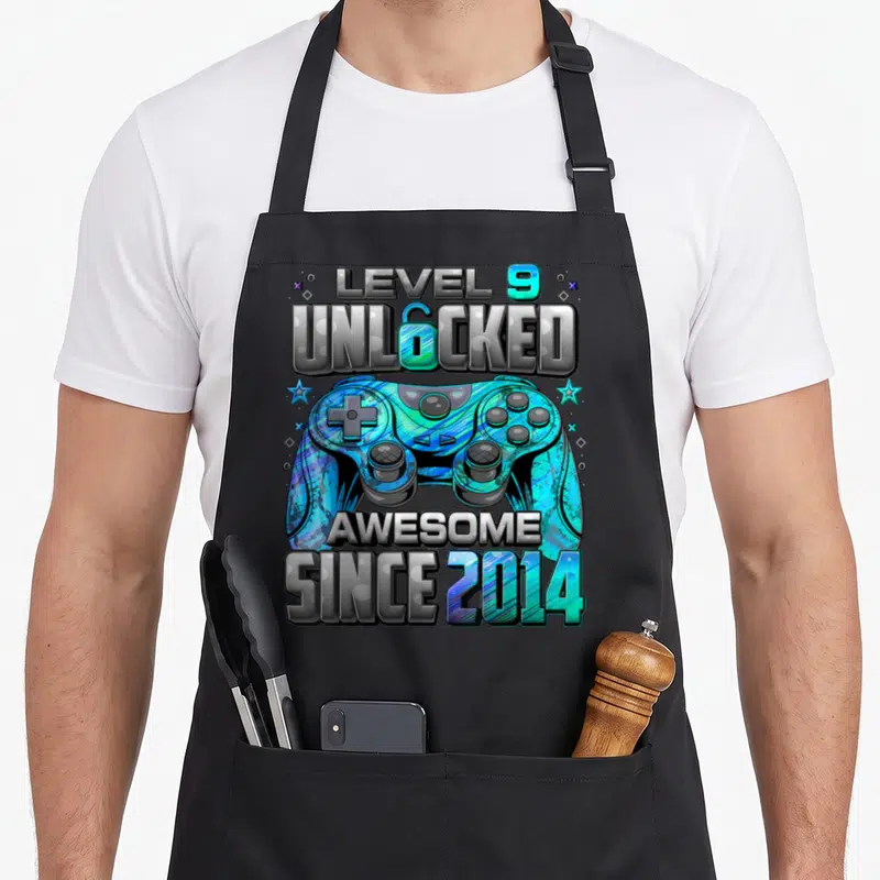 Unlocked 9th gaming aprons - Game controller with stars and text reading Level 9 Unlocked Awesome Since 2014 in grey — shown on a black cotton canvas apron against a white background