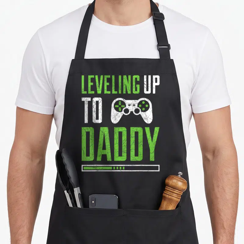 Leveling daddy apron - Green and white text reading LEVELING UP TO DADDY with a game controller and loading bar — shown on a black cotton canvas apron against a white background