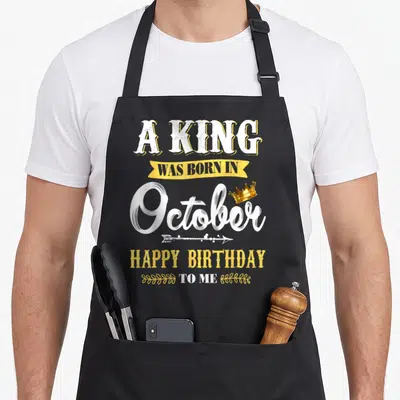 October king apron - Stacked white and yellow text reading A King Was Born In October Happy Birthday To Me with gold — shown on a black cotton canvas apron against a white background
