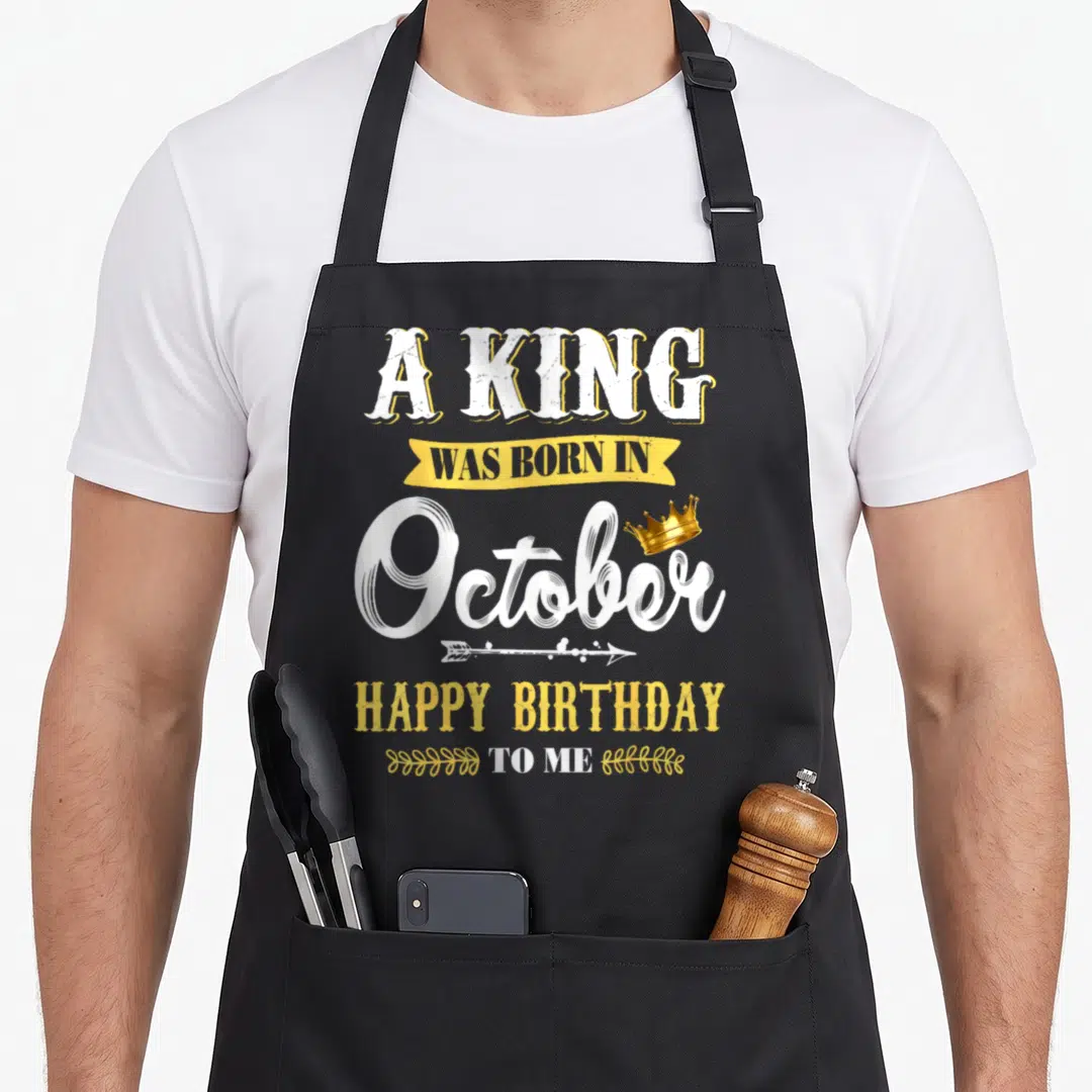 October king apron - Stacked white and yellow text reading A King Was Born In October Happy Birthday To Me with gold — shown on a black cotton canvas apron against a white background