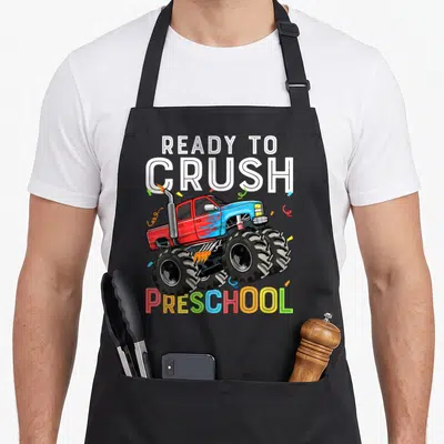 Pre ready truck apron - Monster truck illustration with confetti and colorful PRESCHOOL text — shown on a black cotton canvas apron against a white background