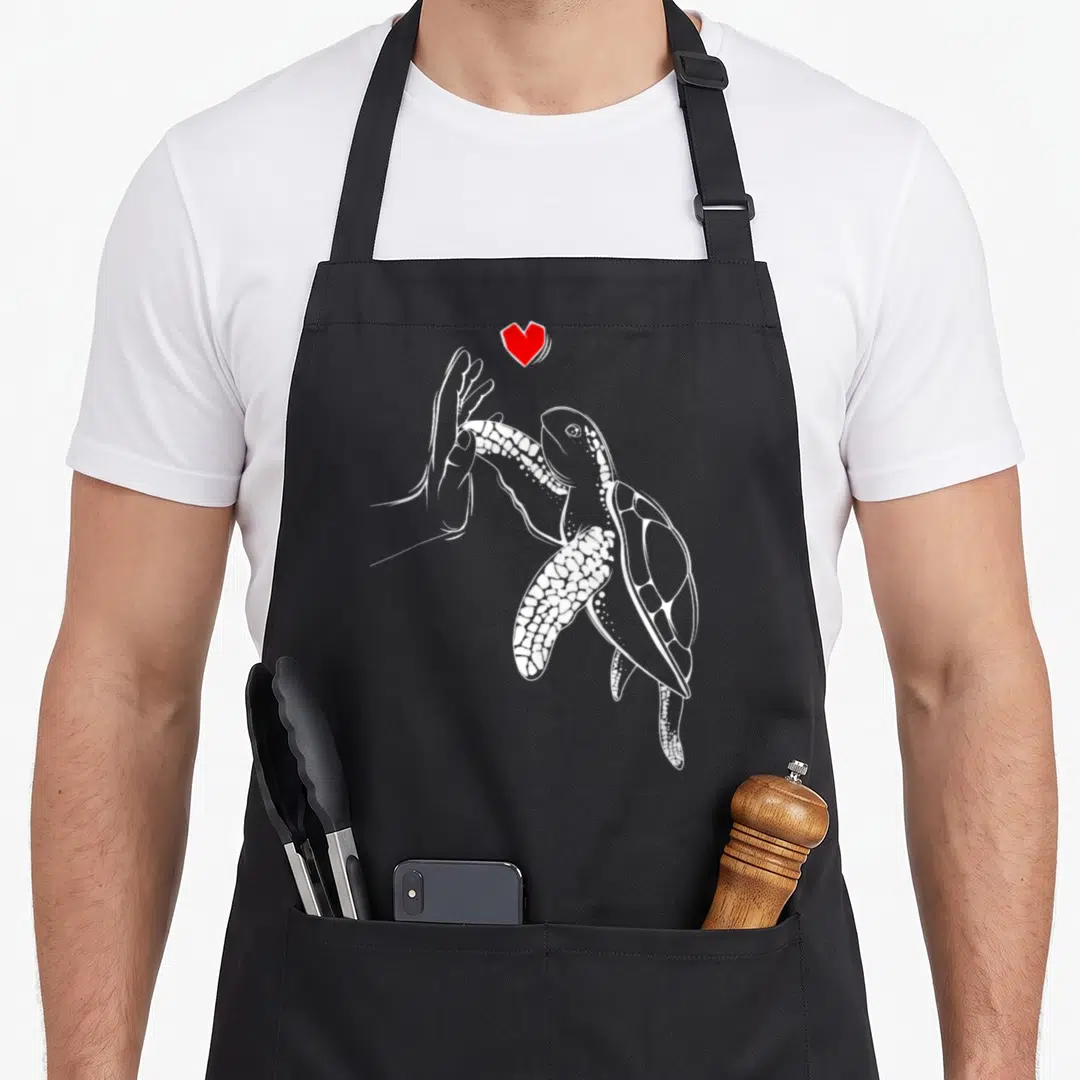 Vintage turtle lover apron - White sea turtle facing left touching human hand with red heart above — shown on a black cotton canvas apron against a white background