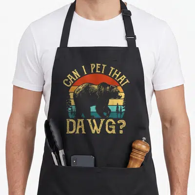 Dawg funny dog apron - Retro bear silhouette against red orange yellow blue sunset with arched text CAN I PET THAT DAWG — shown on a black cotton canvas apron against a white background