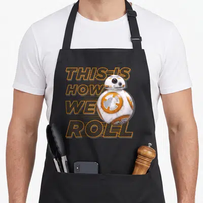 Roll worlds greatest apron - Stacked orange text reading THIS IS HOW WE ROLL next to a white and orange spherical droid — shown on a black cotton canvas apron against a white background