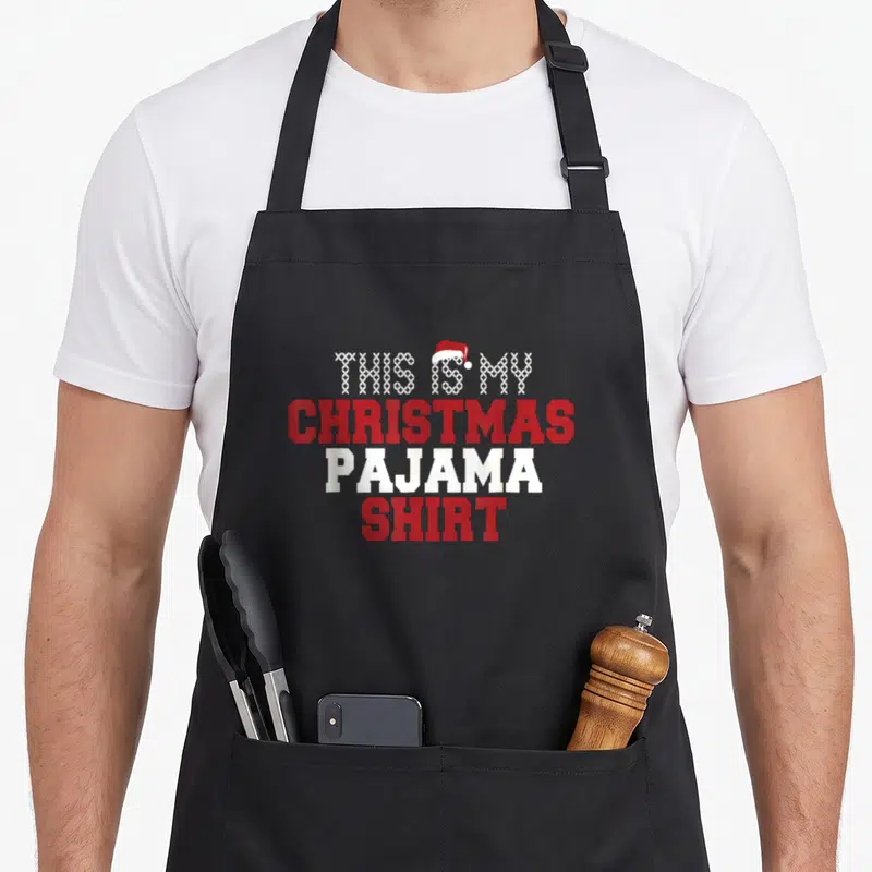 Pajama women's christmas apron - Stacked red and white text reading THIS IS MY CHRISTMAS PAJAMA SHIRT with a Santa hat — shown on a black cotton canvas apron against a white background
