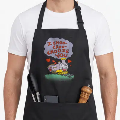 Choose valentine's day apron - Cartoon train with thought bubble reading I Choo Choo Choose You and scattered red hearts — shown on a black cotton canvas apron against a white background